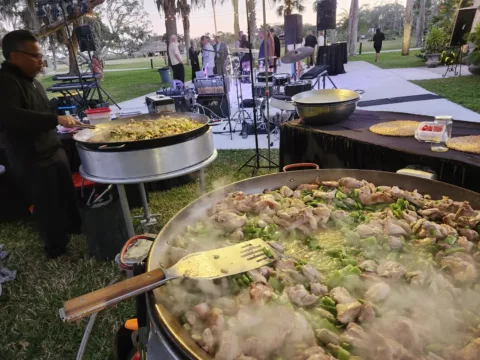 Chef cooking chicken and green bean paella outdoors at an evening party