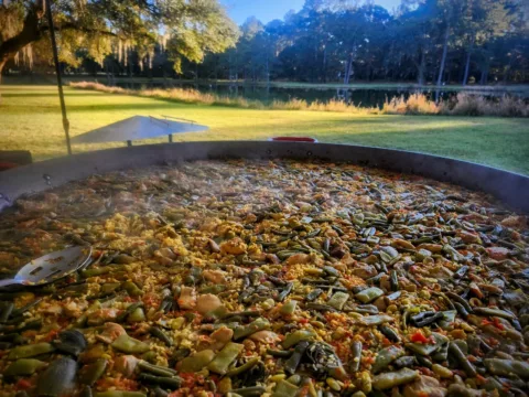 Pan of vegetable and seafood paella cooking on a lawn by a lake