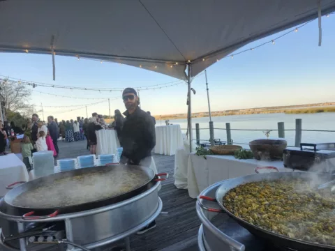 Chef tending paella pans under a tent at a sunset waterfront reception